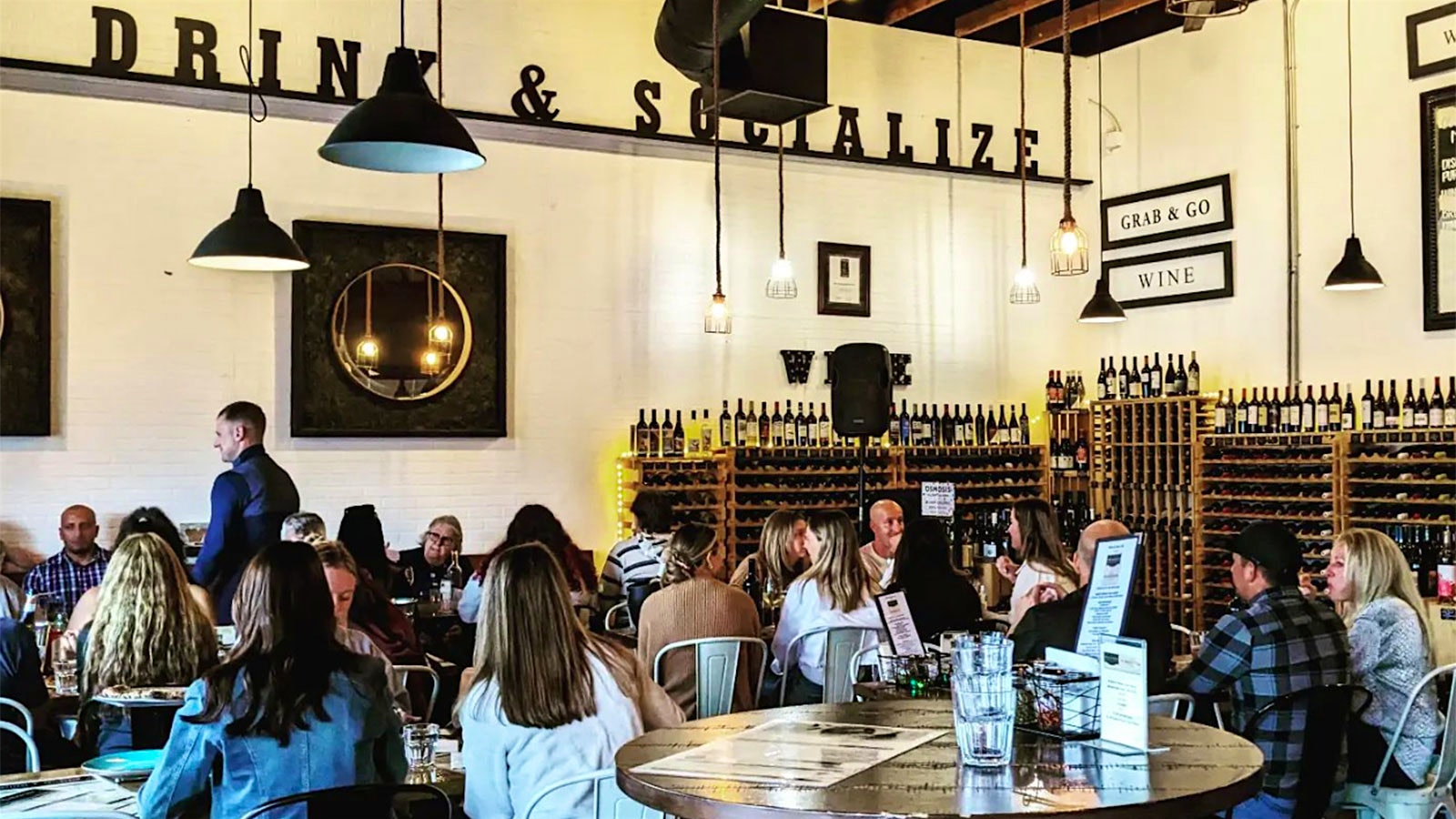 Diners sitting in the dining room at The Privateer Coal Fire Pizza, where there are wood racks of wine bottles, metal chairs and a sign on the wall that says “Drink and Socialize”
